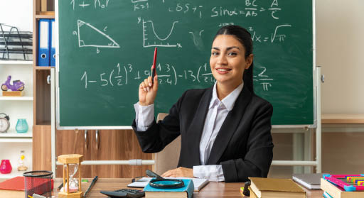 impressed young female teacher sits at table with school tools raising pen in classroom