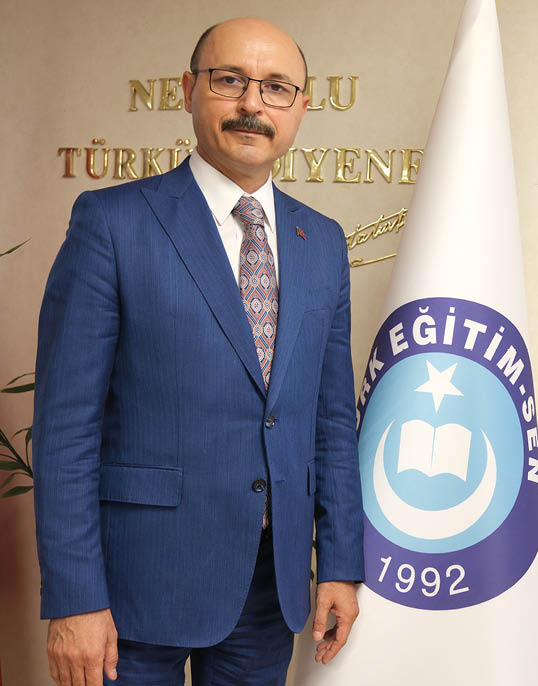 A man wearing a blue suit and a tie stands in front of a white and blue flag. Description generated by AI