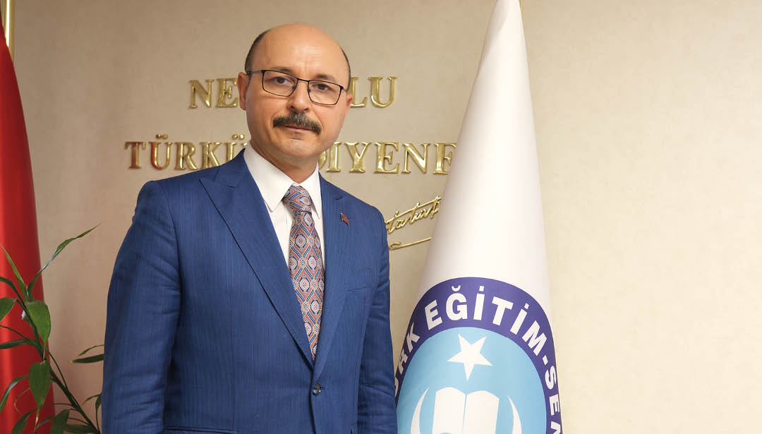 A man wearing a blue suit and a tie stands in front of a white and blue flag. Description generated by AI
