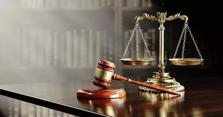 A wooden judge's gavel rests on a polished surface next to a brass balance scale with a blurred bookshelf background. Description generated by AI