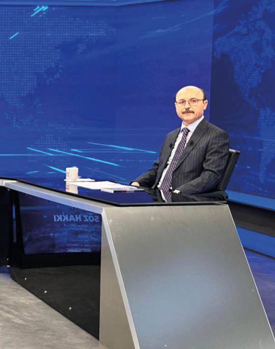 Two people in suits sit at opposite ends of a modern curved news desk with a blue digital world map background. Description generated by AI