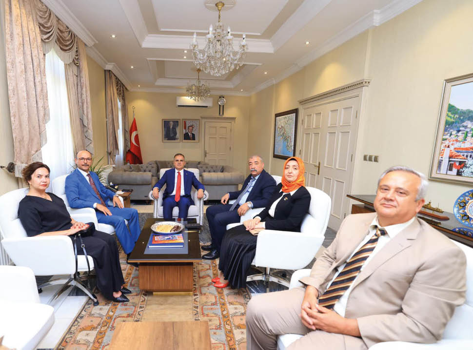 Six adults dressed in formal attire are seated in a well-lit, elegant office with a chandelier, Turkish flag, and framed pictures on the walls. Description generated by AI