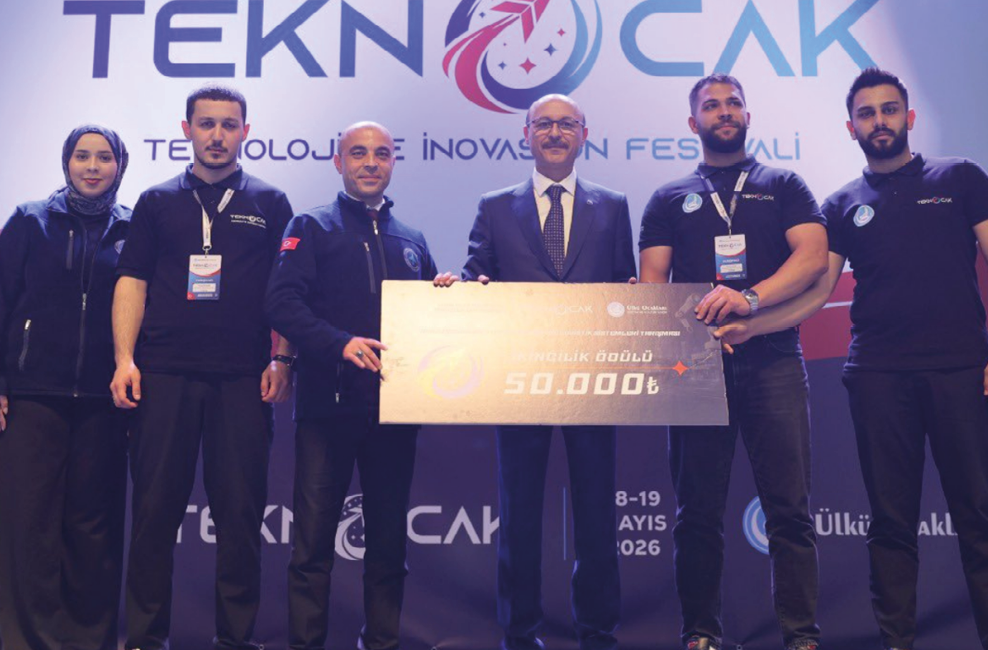 Six adults stand on a stage holding a large check with a technology festival backdrop behind them. Description generated by AI
