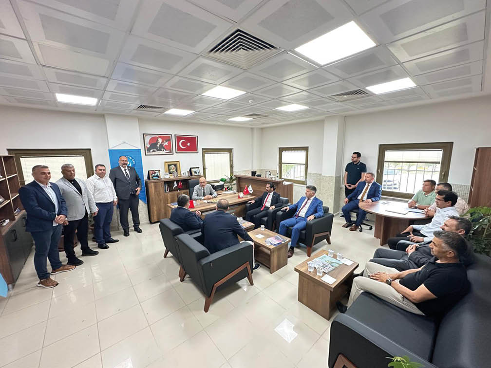 A group of people dressed in formal and casual attire are gathered in a well-lit office with modern furniture, engaged in a meeting or discussion. Description generated by AI