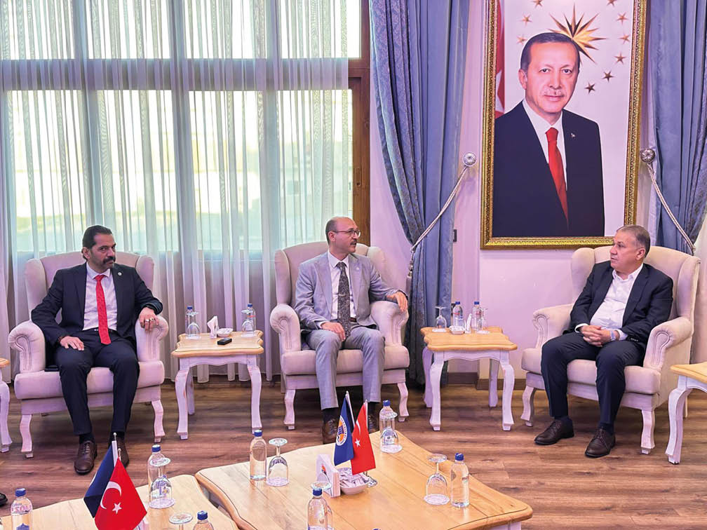 Three people in formal suits are seated in armchairs in a room with wooden floors, small tables with water bottles, and a large framed portrait on the wall behind them. Description generated by AI