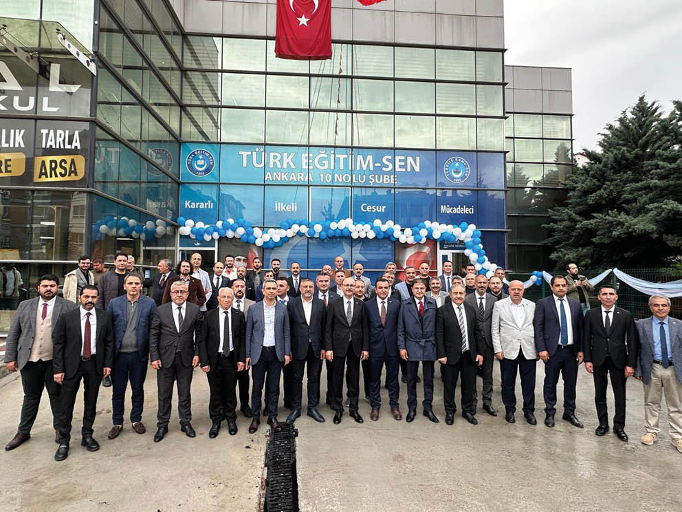 A large group of people dressed in formal attire stands in front of a building decorated with blue and white balloons and a banner reading “T RK E T M-SEN ANKARA 10 NOLU  UBE" under two hanging flags.  Description generated by AI