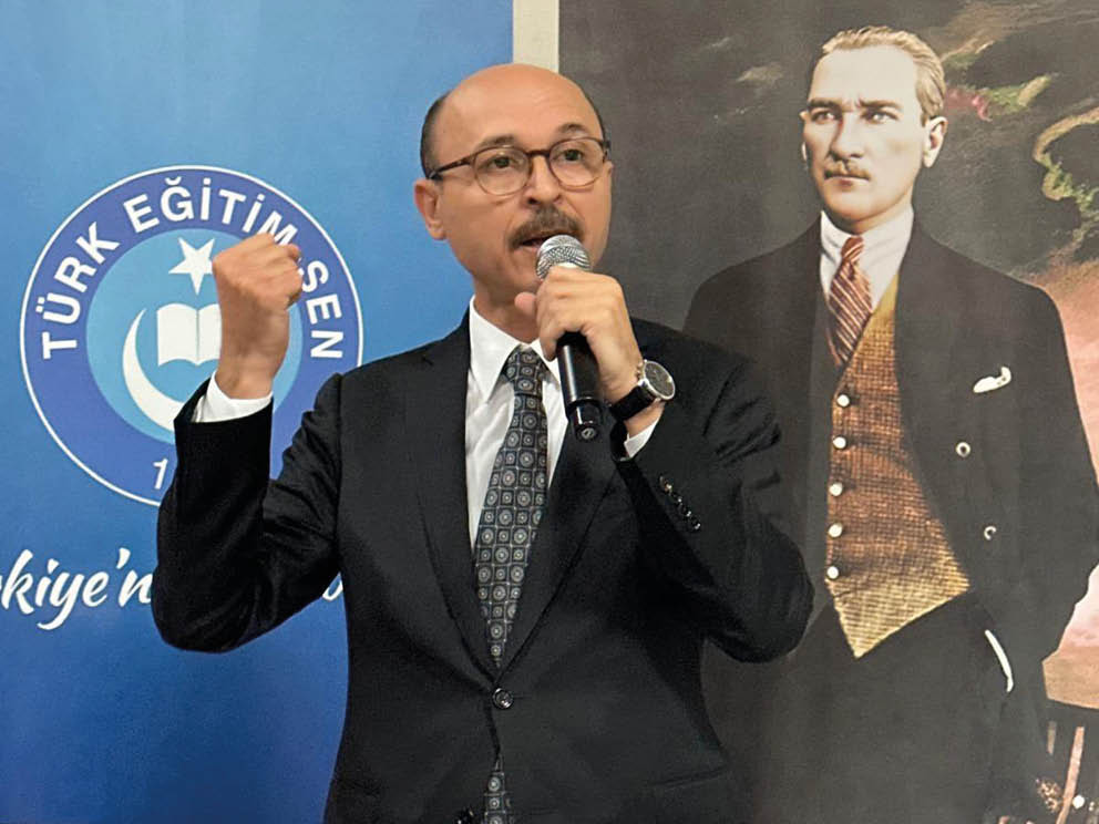 A person in a suit and tie is speaking into a microphone with a raised fist in front of banners displaying the logo and text of T rk E itim-Sen. Description generated by AI