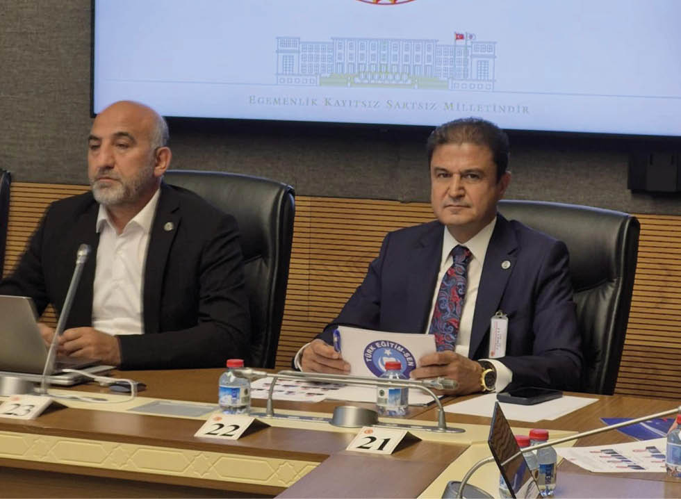 Two people in formal attire sit at a conference table with microphones, documents, and water bottles, beneath a screen displaying the emblem of the Turkish Grand National Assembly. Description generated by AI