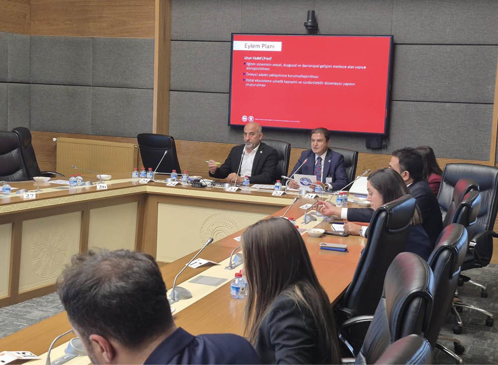 People in business attire are seated around a conference table engaged in a meeting with a presentation displayed on a screen in a modern office setting. Description generated by AI