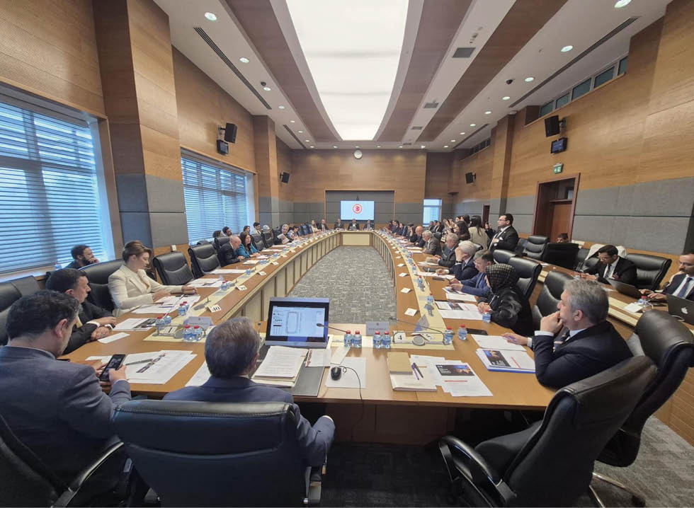 People in business attire are seated around a large U-shaped conference table in a modern meeting room with wood-paneled walls and large windows, engaged in a formal discussion with documents and laptops in front of them. Description generated by AI
