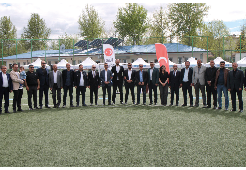 A group of adults dressed in formal and semi-formal attire stand in a line on a grassy field with flags and tents in the background. Description generated by AI