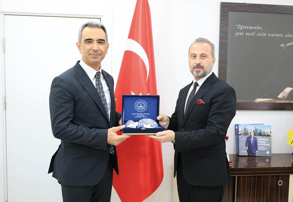Two adults in suits stand in front of a flag while holding a blue box containing decorative items. Description generated by AI