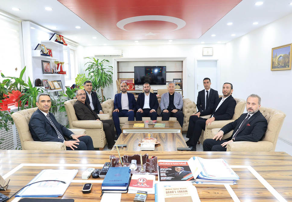 Seven people dressed in formal attire are seated on beige sofas around a wooden coffee table in a well-lit office with plants, shelves, and a television on the wall. Description generated by AI