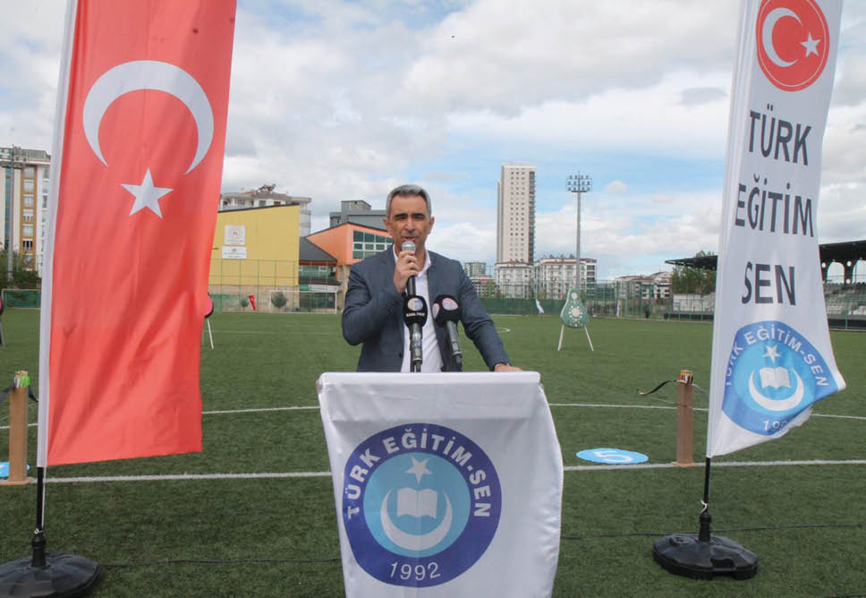 A person in a suit is speaking into a microphone behind a podium with a banner that reads “T RK E T M SEN 1992" on a sports field with two flags displaying the same text on either side.  Description generated by AI