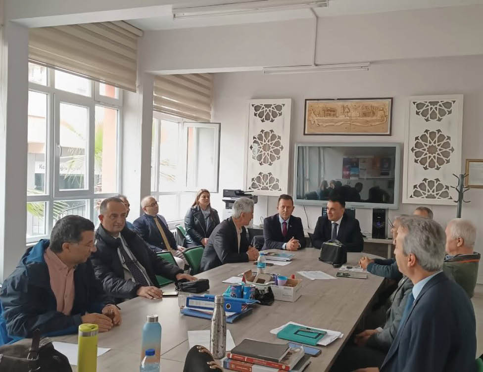 A group of adults in formal and casual clothing are seated around a conference table in a well-lit office with large windows, engaged in a meeting. Description generated by AI