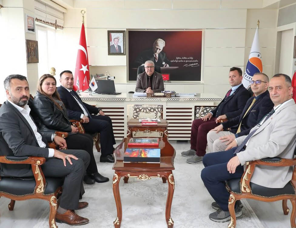 Seven people in formal attire are seated in a meeting room with a person at a desk in the center, Turkish flags and a portrait with a quote are displayed on the wall behind. Description generated by AI