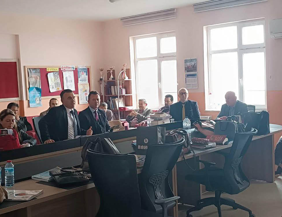 People in formal attire are seated and standing around desks in a well-lit office or meeting room with large windows and various office supplies. Description generated by AI