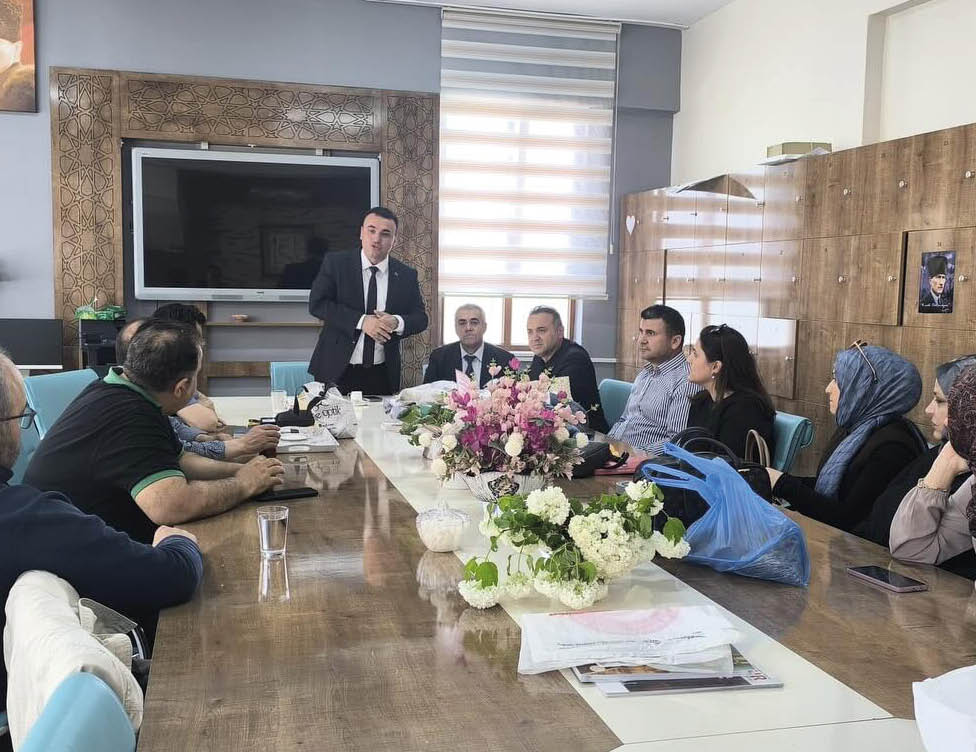 People dressed in formal and casual clothing are seated around a conference table with floral centerpieces while one person stands speaking in a modern office setting. Description generated by AI