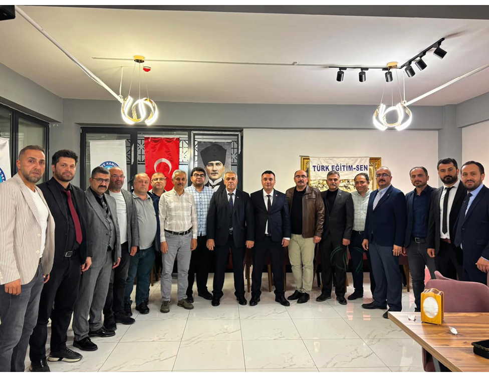 A group of adults dressed in formal and casual clothing stand together indoors in front of flags and a banner with Turkish text. Description generated by AI