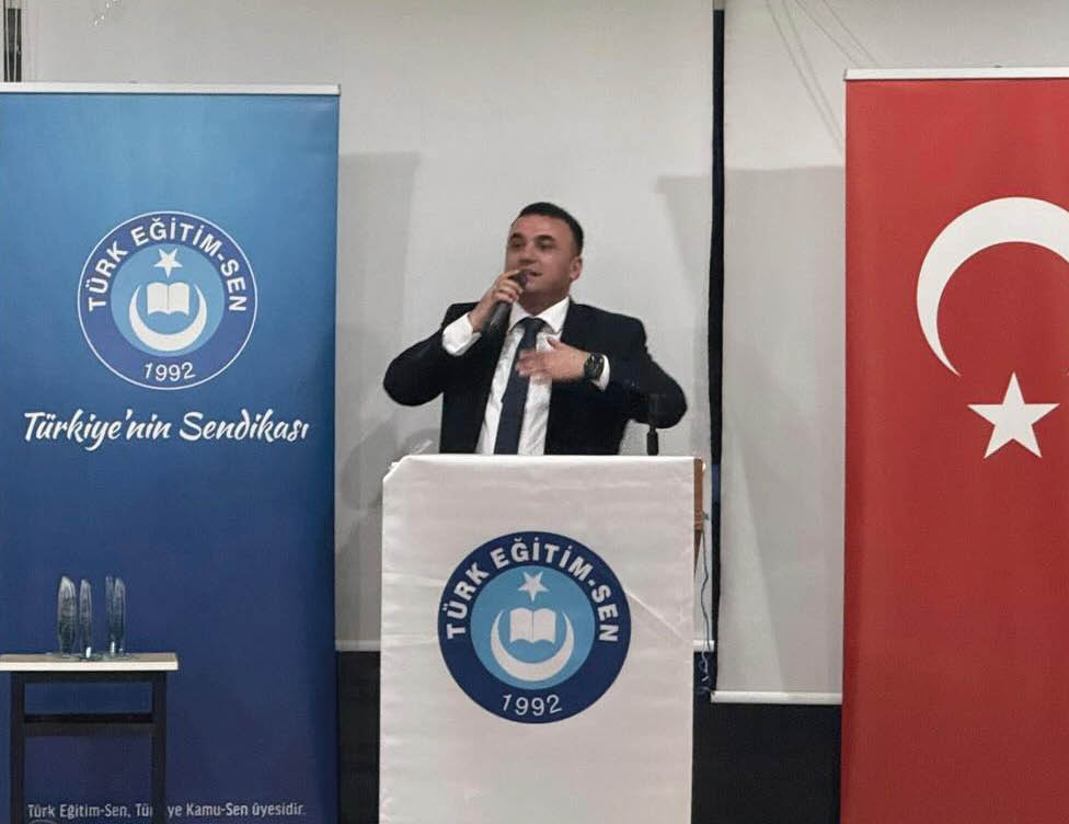 A person in a suit is speaking at a podium with a microphone between two banners, one blue with the text “T RK E T M-SEN 1992 T rkiye'nin Sendikas " and the other red with a white star and crescent.  Description generated by AI