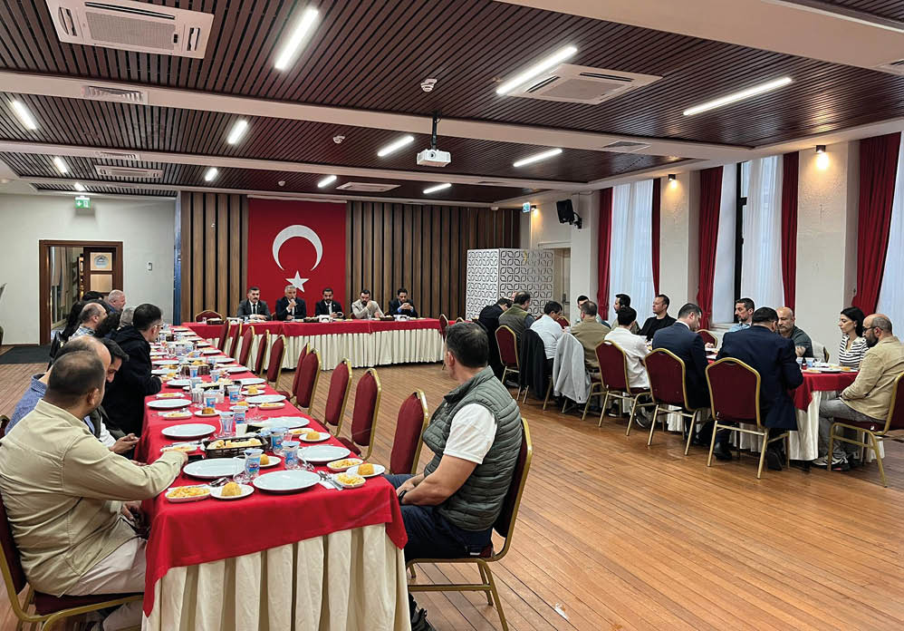 Adults are seated around tables in a meeting room with a large flag featuring a crescent and star hanging on the wall. Description generated by AI