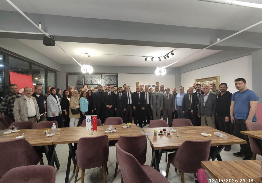 A group of formally dressed people standing closely together in a well-lit room with tables and small flags in the foreground. Description generated by AI