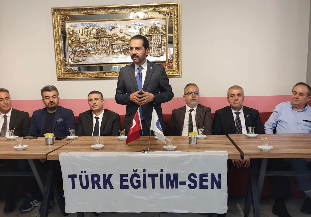 A group of five people in formal attire are seated at a table with two flags and a banner that reads “T RK E T M-SEN" while one person stands and speaks.  Description generated by AI