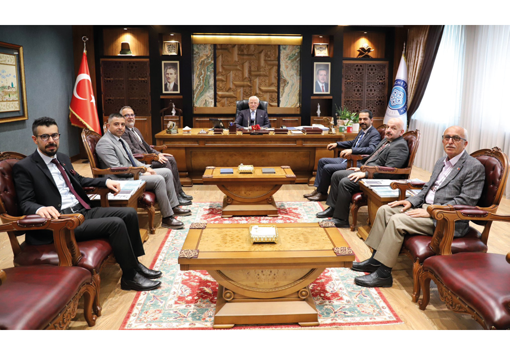 Several people in formal attire are seated around a large wooden desk in an ornate office setting with decorative rugs and plants. Description generated by AI