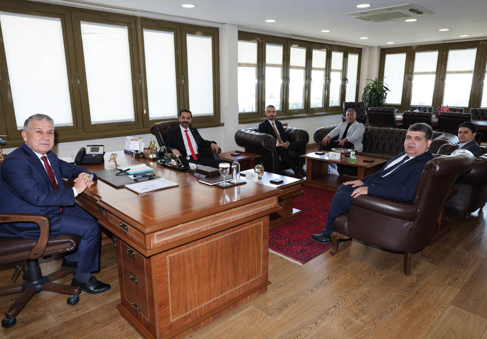 Four adults in formal attire are seated in a well-lit office with large windows, leather chairs, a wooden desk, and a red carpet. Description generated by AI