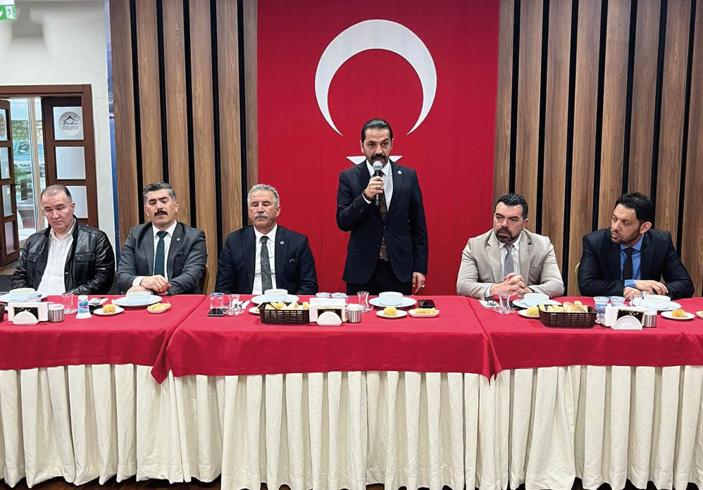 Four adults in formal attire sit at a table with red tablecloths and white skirts while one adult in a dark suit stands speaking into a microphone in front of a large red flag with a white crescent and star. Description generated by AI