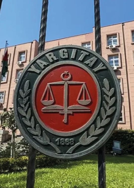 A metal sign with the word “YARGITAY" and a scale symbol is mounted on a fence in front of a multi-story building with several windows and air conditioning units. Description generated by AI