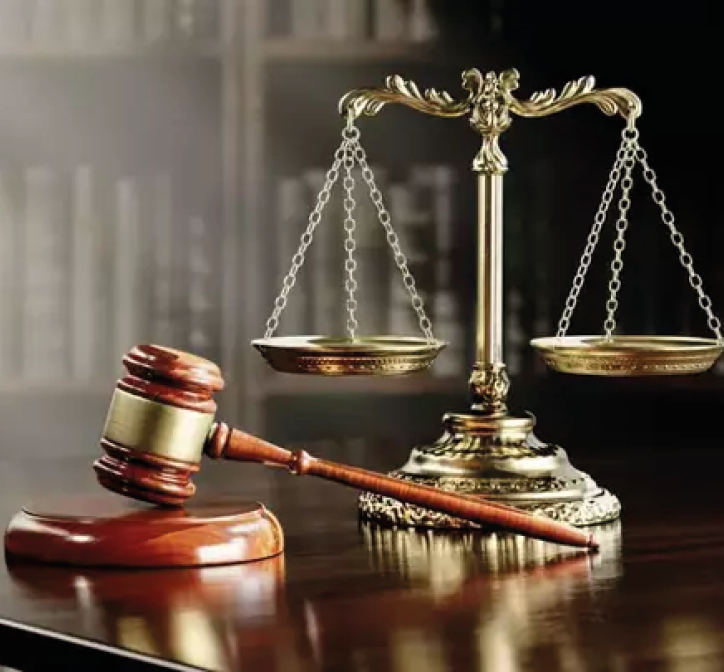 A wooden judge's gavel rests on a polished surface next to a brass balance scale with a blurred bookshelf background. Description generated by AI