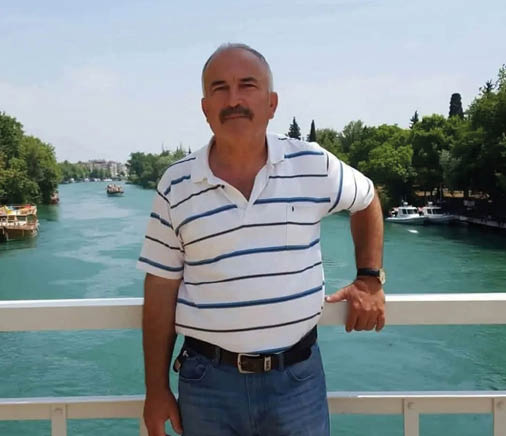 A person wearing a white and blue striped polo shirt and jeans is standing on a bridge over a river with boats and trees in the background. Description generated by AI
