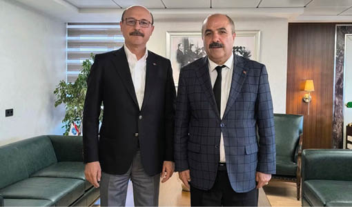 Two adults wearing suits stand side by side in a modern office setting with green sofas and framed pictures on the wall behind them. Description generated by AI