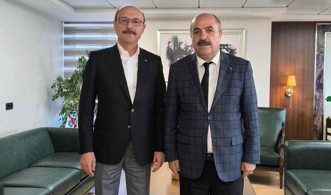 Two adults wearing suits stand side by side in a modern office setting with green sofas and framed pictures on the wall behind them. Description generated by AI