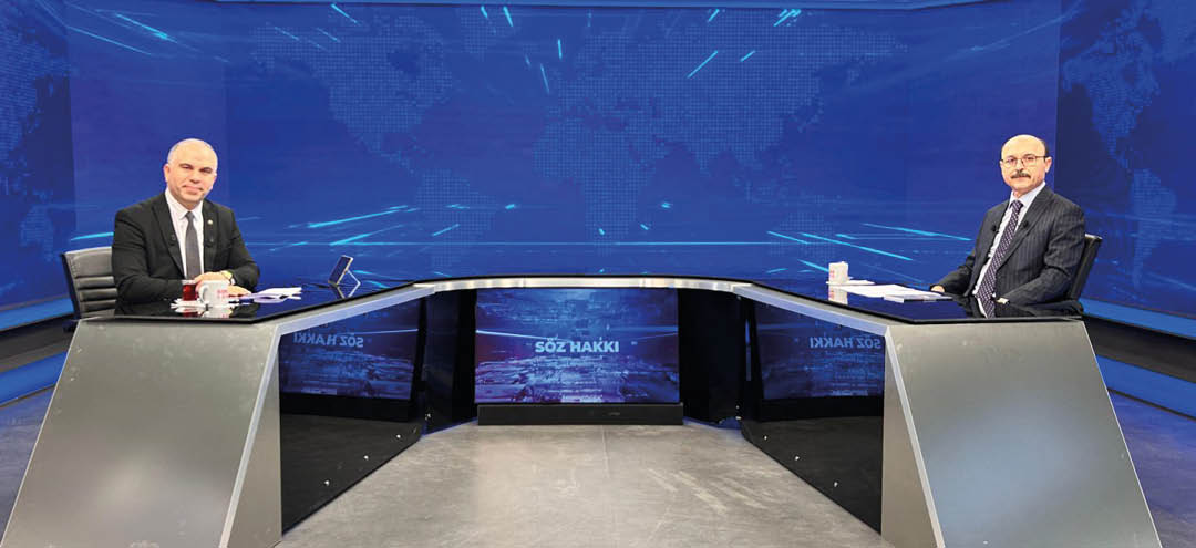 Two people in suits sit at opposite ends of a modern curved news desk with a blue digital world map background. Description generated by AI