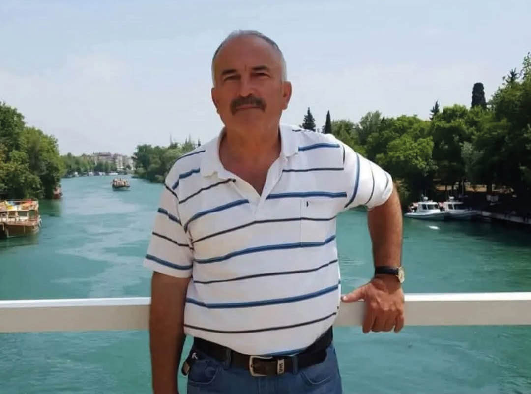 A person wearing a white and blue striped polo shirt and jeans is standing on a bridge over a river with trees and boats in the background. Description generated by AI