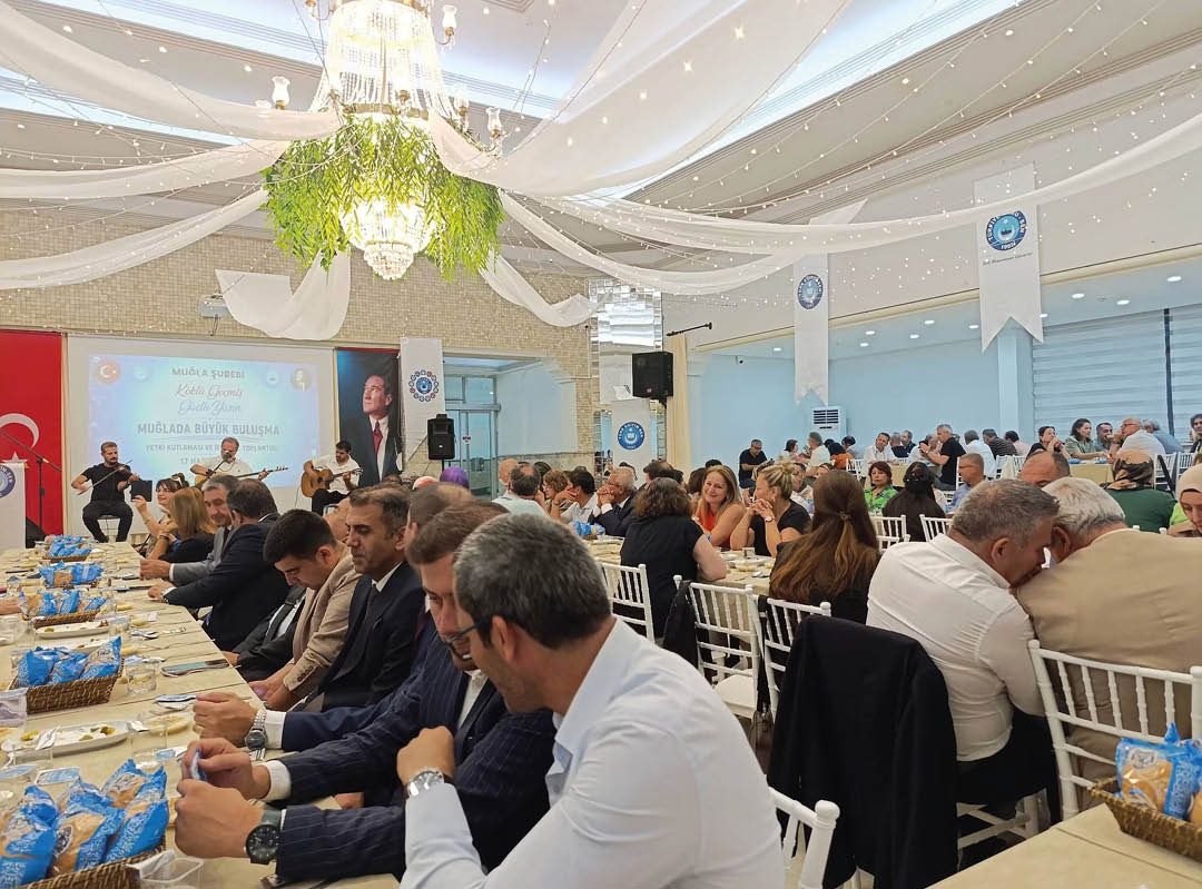 A large group of people seated at long tables in a decorated indoor venue are attentively listening to a speaker near a screen displaying a presentation. Description generated by AI