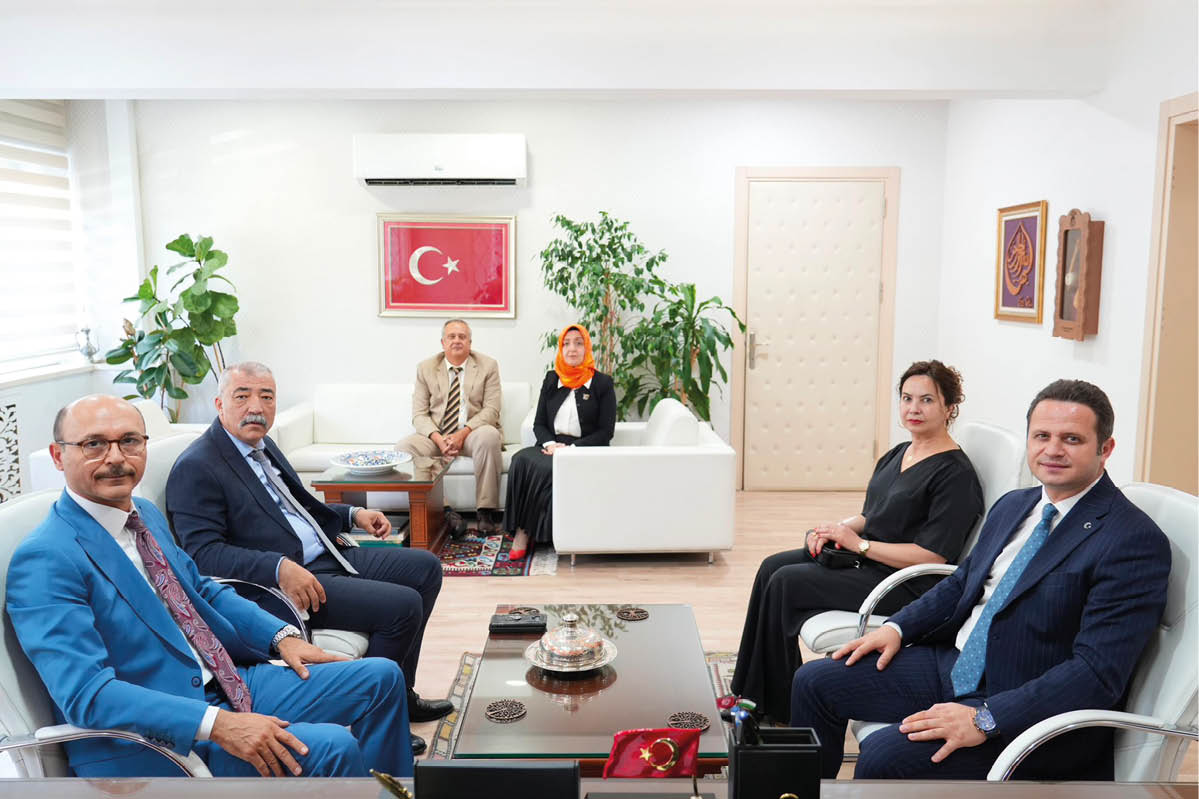 Six adults dressed in formal and traditional clothing are seated in a modern office with plants, a Turkish flag on the wall, and a coffee table with decorative items. Description generated by AI