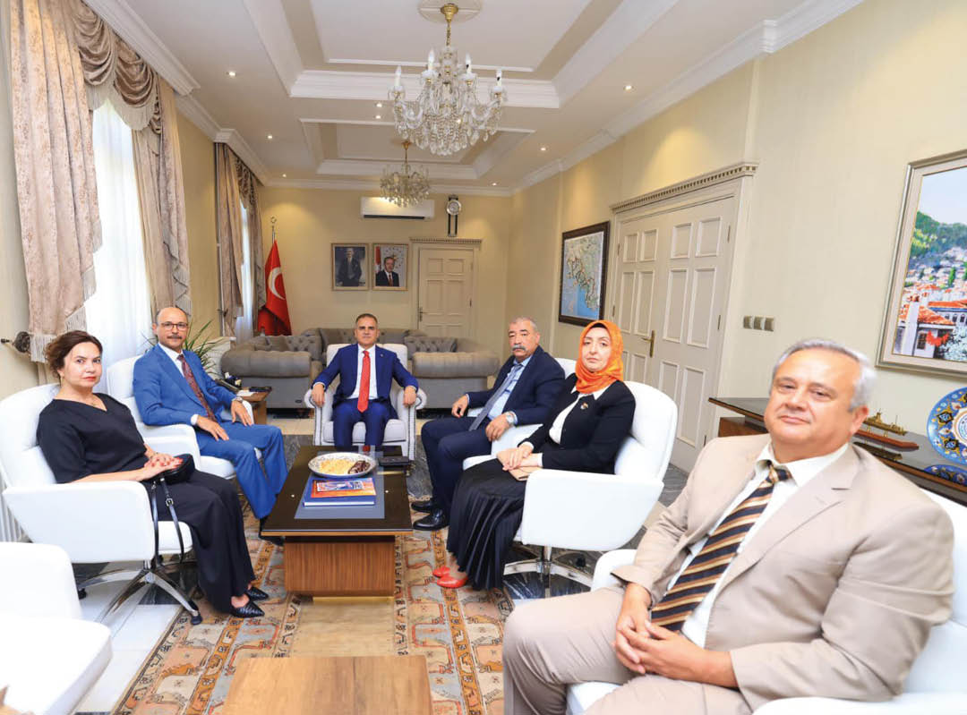 Six adults dressed in formal attire are seated in a well-lit, elegant office with a chandelier, Turkish flag, and framed pictures on the walls. Description generated by AI
