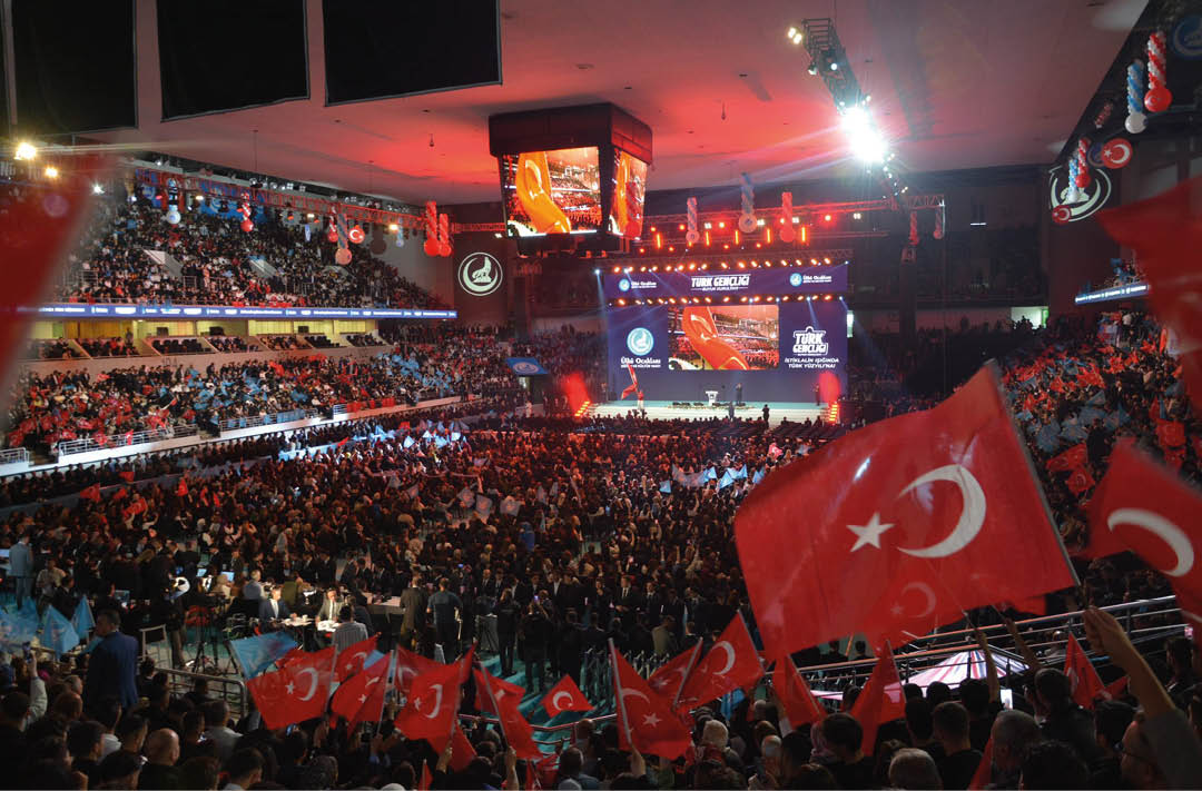 A large crowd of people is gathered indoors at a brightly lit event with a stage, large screens displaying logos and text, and many attendees holding blue flags. Description generated by AI