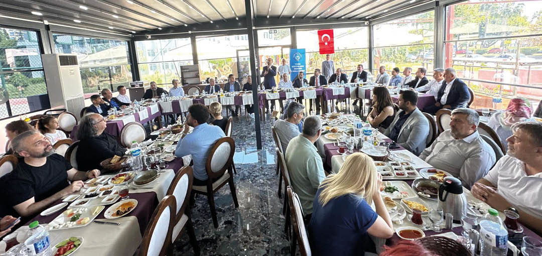 People are seated around a large U-shaped table in a bright, covered outdoor dining area, engaged in a formal meal or meeting with plates of food and drinks in front of them. Description generated by AI