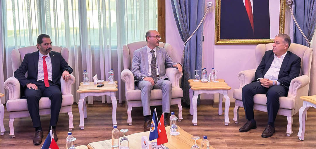 Three people in formal suits are seated in armchairs in a room with wooden floors, small tables with water bottles, and a large framed portrait on the wall behind them. Description generated by AI