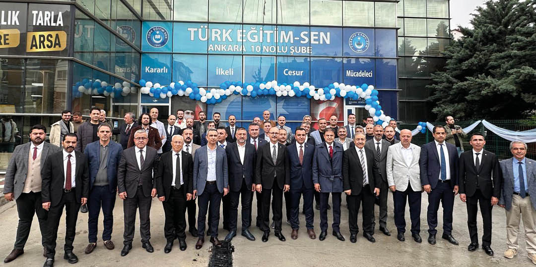 A large group of people dressed in formal attire stands in front of a building decorated with blue and white balloons and a banner reading “T RK E T M-SEN ANKARA 10 NOLU  UBE" under two hanging flags.  Description generated by AI