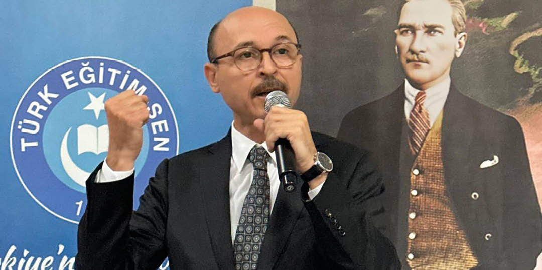 A person in a suit and tie is speaking into a microphone with a raised fist in front of banners displaying the logo and text of T rk E itim-Sen. Description generated by AI