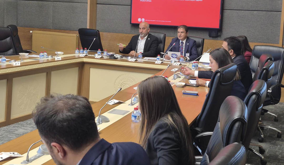 People in business attire are seated around a conference table engaged in a meeting with a presentation displayed on a screen in a modern office setting. Description generated by AI