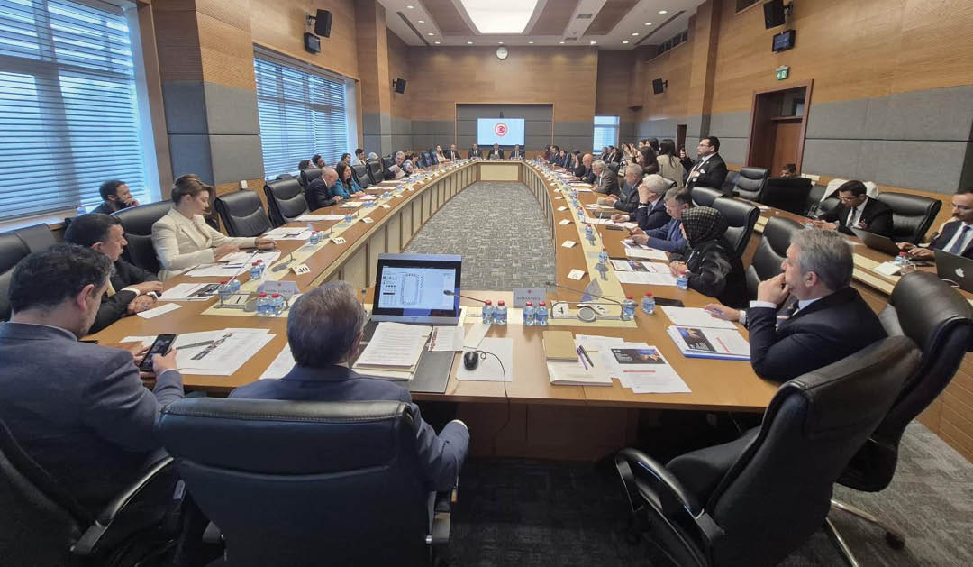 People in business attire are seated around a large U-shaped conference table in a modern meeting room with wood-paneled walls and large windows, engaged in a formal discussion with documents and laptops in front of them. Description generated by AI
