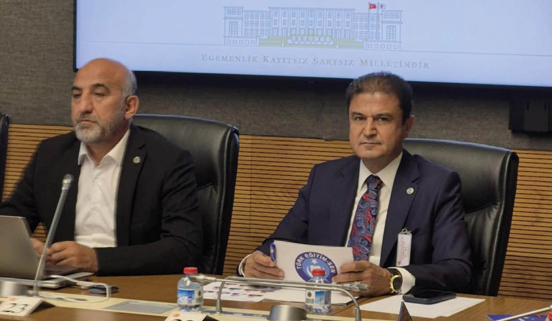 Two people in formal attire sit at a conference table with microphones, documents, and water bottles, beneath a screen displaying the emblem of the Turkish Grand National Assembly. Description generated by AI