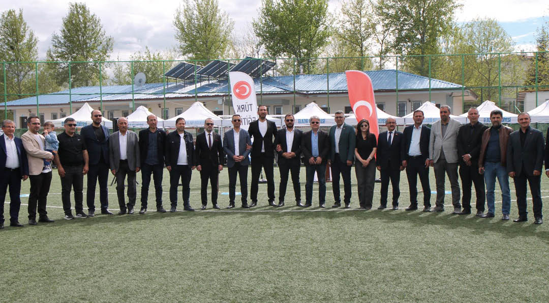 A group of adults dressed in formal and semi-formal attire stand in a line on a grassy field with flags and tents in the background. Description generated by AI