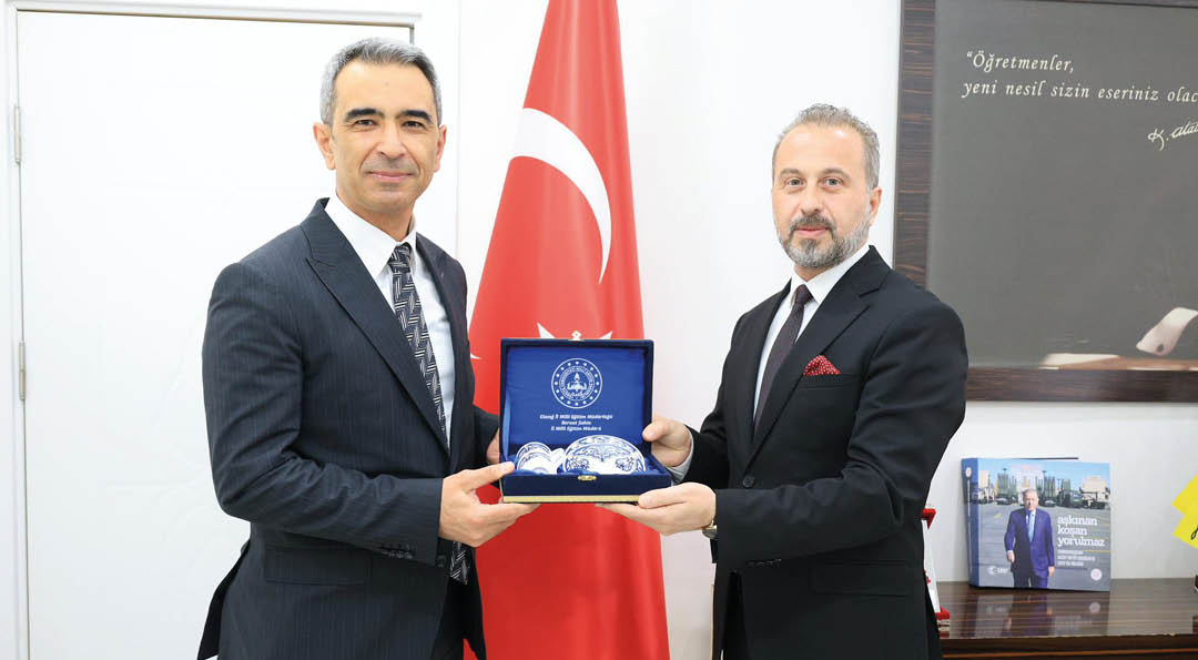 Two adults in suits stand in front of a flag while holding a blue box containing decorative items. Description generated by AI